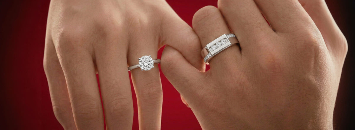 A close-up of two hands wearing rings against a dark red background. The left hand wears a silver engagement ring with a round-cut diamond, and the right hand wears a matching silver band with small diamonds.